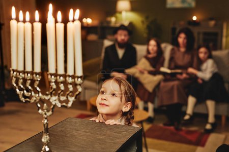 Foto de Retrato de niña judía mirando la vela menorah asombrada mientras celebra Hanukkah con la familia, espacio para copiar - Imagen libre de derechos