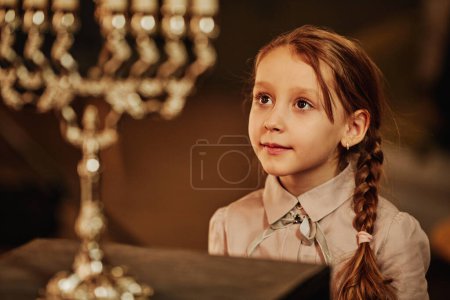 Téléchargez les photos : Portrait de jolie fille juive regardant la bougie menorah pendant les célébrations de Hanoukka - en image libre de droit