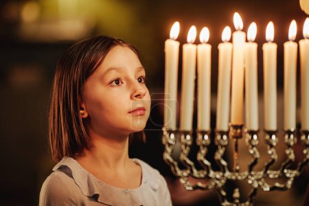 Téléchargez les photos : Vue de côté portrait de jeune fille regardant la bougie menorah pendant les célébrations de Hanoukka - en image libre de droit