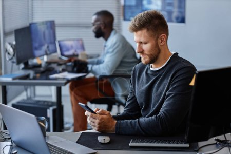Photo for Side view portrait of young Caucasian man using smartphone at workplace while working in mobile software development, copy space - Royalty Free Image