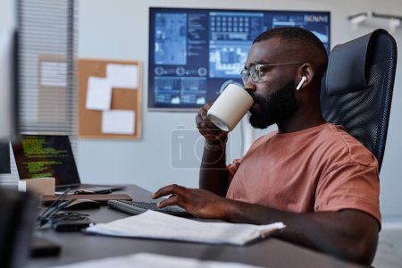 Photo for Side view portrait of black man drinking coffee at workplace while working in IT software development - Royalty Free Image