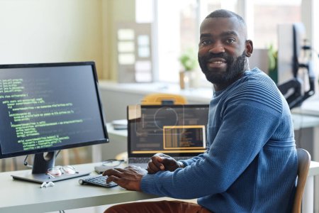 Foto de Retrato del hombre negro como programador de computadoras mirando la cámara mientras escribe código en el lugar de trabajo de la oficina - Imagen libre de derechos