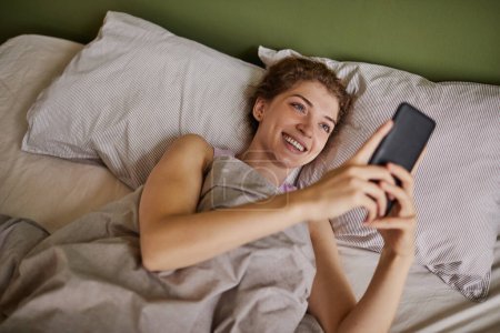 Photo for Young happy woman reading message in her smartphone and smiling while resting in her bed during weekends - Royalty Free Image