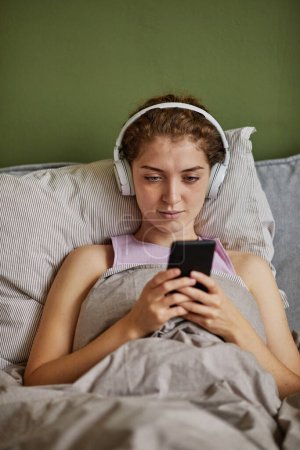 Foto de Chica joven en auriculares inalámbricos escuchando audiolibros en su teléfono inteligente mientras está acostada en la cama en el dormitorio - Imagen libre de derechos