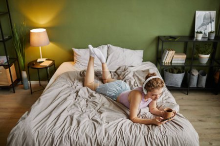 Téléchargez les photos : Vue grand angle de la jeune femme dans les écouteurs en utilisant le téléphone portable tout en étant couché sur son lit avec une tasse de café - en image libre de droit