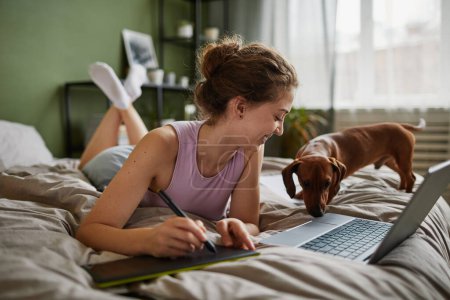 Foto de Chica dibujo en la tableta gráfica mientras está acostado en la cama con el ordenador portátil y su perro en el dormitorio en casa - Imagen libre de derechos