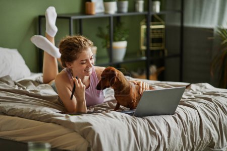 Foto de Chica joven usando el ordenador portátil y jugando con su perro mientras se relaja en la cama durante el tiempo libre en casa - Imagen libre de derechos