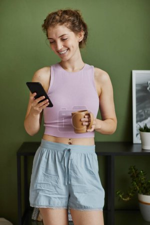 Téléchargez les photos : Jeune femme heureuse recevant un message sur son smartphone tout en buvant du café le matin dans la chambre - en image libre de droit