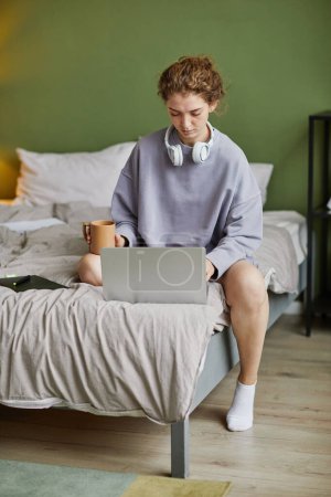 Téléchargez les photos : Jeune femme assise sur le lit avec une tasse de café et tapant sur ordinateur portable à la maison - en image libre de droit