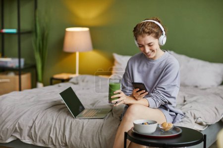 Téléchargez les photos : Jeune femme dans les écouteurs sans fil boire smoothie et en utilisant le téléphone portable pendant le petit déjeuner dans la chambre - en image libre de droit