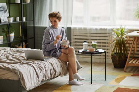 Téléchargez les photos : Jeune femme prenant le petit déjeuner et utilisant un ordinateur portable tout en étant assis sur le lit dans la chambre à la maison - en image libre de droit
