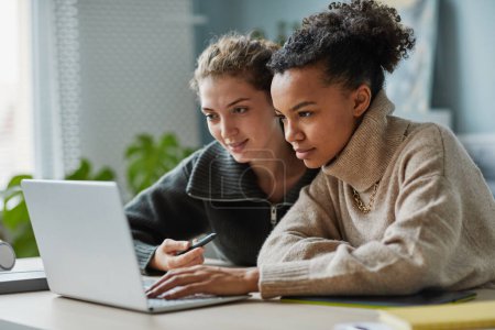 Téléchargez les photos : Deux jeunes collègues assis à table devant un ordinateur portable et regardant l'écran pendant un appel vidéo - en image libre de droit