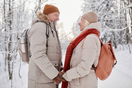 Photo for Side view portrait of happy senior couple enjoying walk in winter forest and holding hands - Royalty Free Image