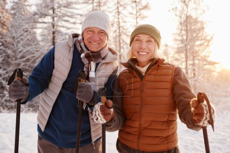 Photo for Waist up portrait of active senior couple enjoying Nordic walk with poles in winter forest and looking at camera - Royalty Free Image