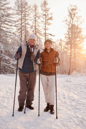 Photo for Vertical portrait of active senior couple enjoying Nordic walk with poles in winter forest and looking at camera - Royalty Free Image