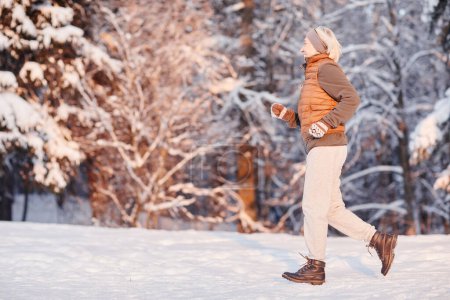Téléchargez les photos : Portrait latéral de femme mûre sportive courant dans la forêt d'hiver éclairée par la lumière du coucher du soleil, espace de copie - en image libre de droit