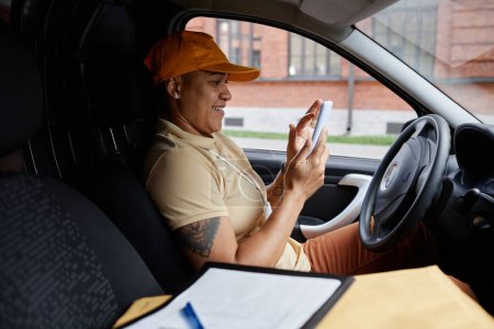 Photo for Side view portrait of female delivery driver using smartphone app in van - Royalty Free Image