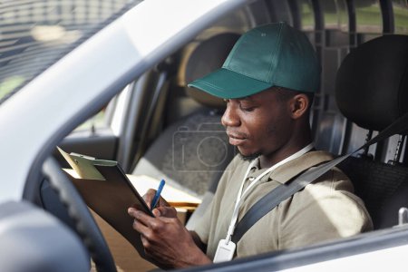 Téléchargez les photos : Portrait latéral de jeune homme noir écrivant sur presse-papiers dans la camionnette de livraison, espace de copie - en image libre de droit