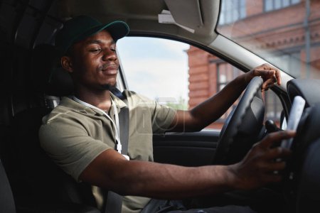 Foto de Retrato de vista lateral del joven afroamericano conduciendo furgoneta de reparto y vistiendo uniforme verde, espacio para copiar - Imagen libre de derechos