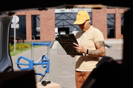 Foto de Retrato del repartidor multiétnico rellenando formularios mientras está parado junto a la camioneta a la luz del sol, espacio para copiar - Imagen libre de derechos