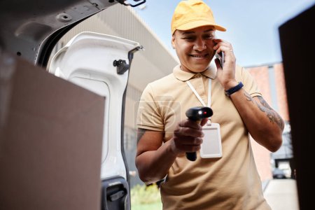 Foto de Retrato del trabajador de reparto sonriente hablando por teléfono inteligente y códigos de escaneo en paquetes en furgoneta - Imagen libre de derechos