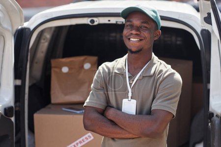 Photo for Waist up portrait of black young man as delivery man standing with arms crossed by van in sunlight and smiling at camera - Royalty Free Image
