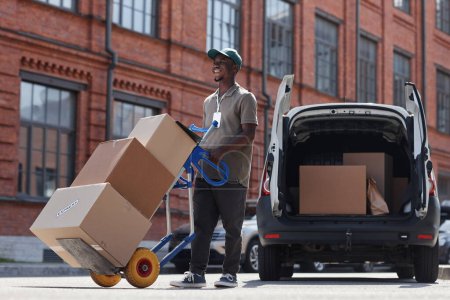 Photo for Full length portrait of smiling delivery worker unloading boxes outdoors in sunlight, copy space - Royalty Free Image
