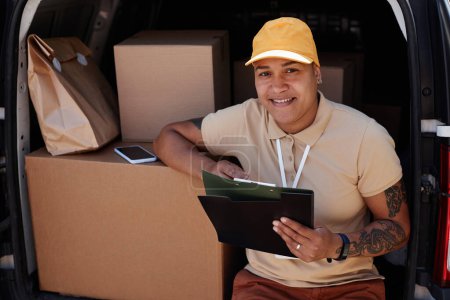 Photo for Portrait of smiling woman as delivery worker looking at camera while writing on clipboard in van trunk, copy space - Royalty Free Image