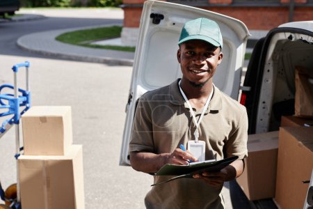 Photo for Waist up portrait of delivery man checking documents and looking at camera while unloading van with packages - Royalty Free Image