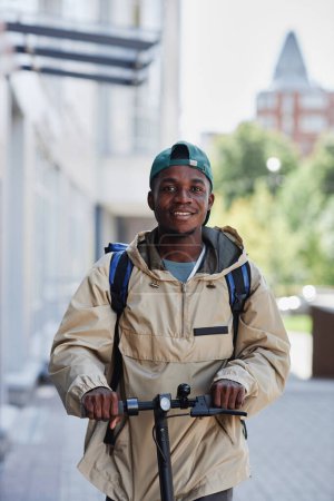 Téléchargez les photos : Portrait vertical de sourire livreur de nourriture garçon équitation scooter électrique dans la rue de la ville - en image libre de droit