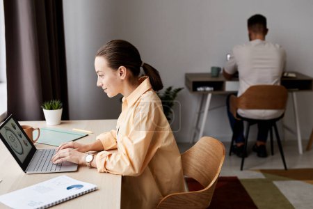 Téléchargez les photos : Vue latérale de la jeune femme d'affaires brune confiante regardant les données graphiques sur l'écran d'ordinateur portable tout en étant assise par le lieu de travail à la maison - en image libre de droit