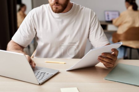 Téléchargez les photos : Gros plan d'un jeune homme en t-shirt blanc tenant un copybook et utilisant un ordinateur portable assis à son bureau pendant une leçon en ligne avec un tuteur - en image libre de droit