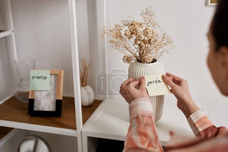 Foto de Enfoque selectivo en las manos de una joven mujer pegando un bloc de notas con una nueva palabra en español en un jarrón blanco con flores secas de pie en el estante - Imagen libre de derechos