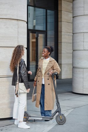 Foto de Retrato vertical de una joven negra con scooter eléctrico hablando con un amigo en el entorno de la ciudad - Imagen libre de derechos