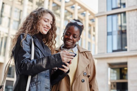 Foto de Retrato de la cintura hacia arriba de dos mujeres jóvenes sonrientes usando un teléfono inteligente mientras están de pie fuera del centro comercial, afroamericanos y caucásicos - Imagen libre de derechos