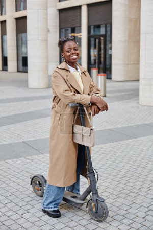 Foto de Retrato vertical de longitud completa de una joven mujer negra montando scooter eléctrico en la ciudad y sonriendo alegremente a la cámara mientras usa gabardina larga - Imagen libre de derechos