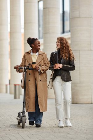 Téléchargez les photos : Portrait vertical pleine longueur de deux jeunes femmes avec scooter électrique marchant vers la caméra en ville - en image libre de droit