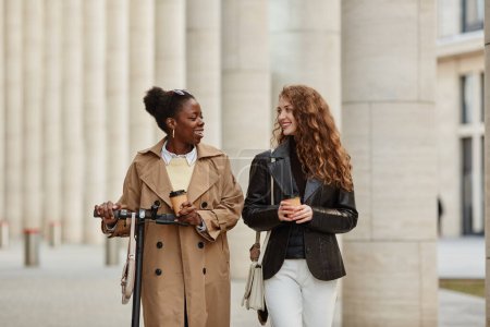 Foto de Retrato de cintura hacia arriba de dos mujeres jóvenes con scooter eléctrico caminando hacia la cámara en la ciudad - Imagen libre de derechos