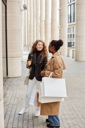 Foto de Retrato vertical de dos jóvenes alegres con bolsas de compras charlando fuera del centro comercial en la ciudad - Imagen libre de derechos