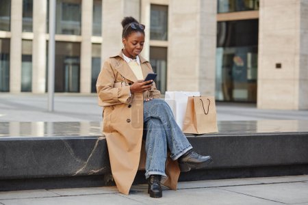 Foto de Retrato de larga duración de una joven negra usando un teléfono inteligente mientras se relaja en el centro de la ciudad después del día de compras - Imagen libre de derechos
