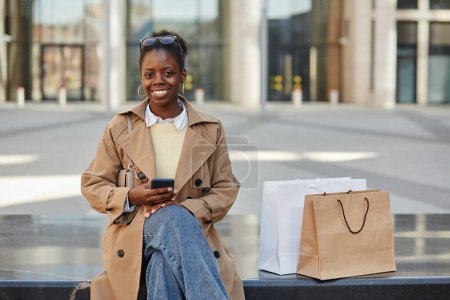 Foto de Retrato de una joven negra usando un teléfono inteligente en el centro de la ciudad y sonriendo a la cámara después del día de compras, espacio para copiar - Imagen libre de derechos