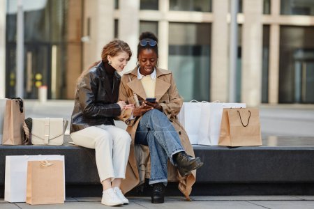 Foto de Retrato de larga duración de dos mujeres jóvenes usando un teléfono inteligente mientras se relaja en el centro de la ciudad después del día de compras - Imagen libre de derechos