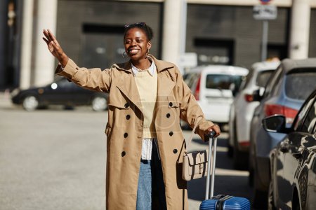 Téléchargez les photos : Portrait de jeune femme noire avec valise saluant taxi dans la rue de la ville et souriant - en image libre de droit