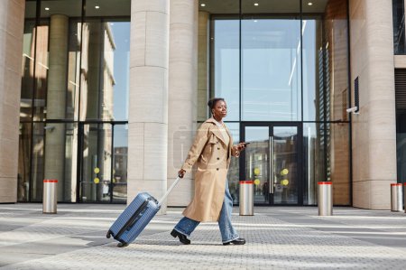 Foto de Retrato de vista lateral de una joven mujer negra con maleta caminando al aire libre en la ciudad por aeropuerto o estación de tren - Imagen libre de derechos