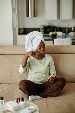 Téléchargez les photos : Portrait vertical d'une jeune femme noire aux cheveux enveloppés dans une serviette jouissant d'une journée d'autosoin à la maison - en image libre de droit