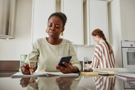 Foto de Retrato de una joven mujer negra usando un teléfono inteligente mientras trabaja desde casa en un entorno de cocina mínimo, espacio para copiar - Imagen libre de derechos