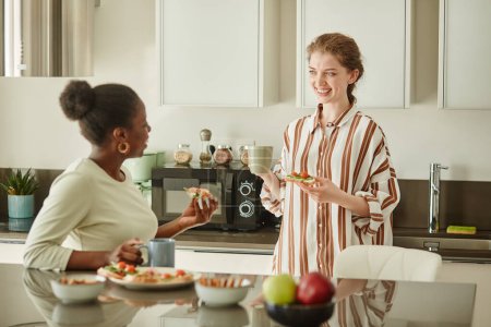 Foto de Retrato de dos mujeres jóvenes charlando alegremente en la acogedora cocina casera por la mañana y disfrutando del desayuno - Imagen libre de derechos