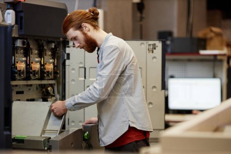 Photo for Side view portrait of bearded young man working in print shop and setting up machines, copy space - Royalty Free Image