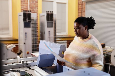 Foto de Retrato de vista lateral de la joven mujer negra operando la máquina de impresión y sosteniendo la hoja de prueba de color, espacio de copia - Imagen libre de derechos