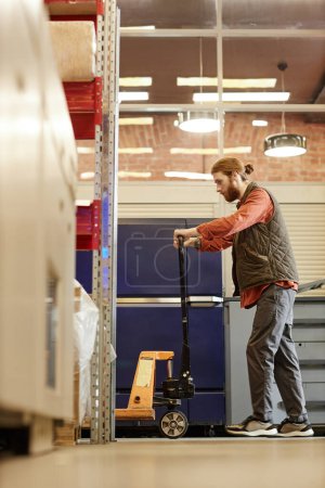 Téléchargez les photos : Vue latérale verticale du travailleur masculin utilisant un cric à palette lors du déplacement de boîtes à l'entrepôt d'usine - en image libre de droit